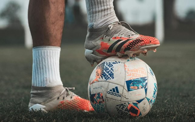 Close-up of a soccer player wearing Adidas cleats, holding a branded soccer ball on grass.